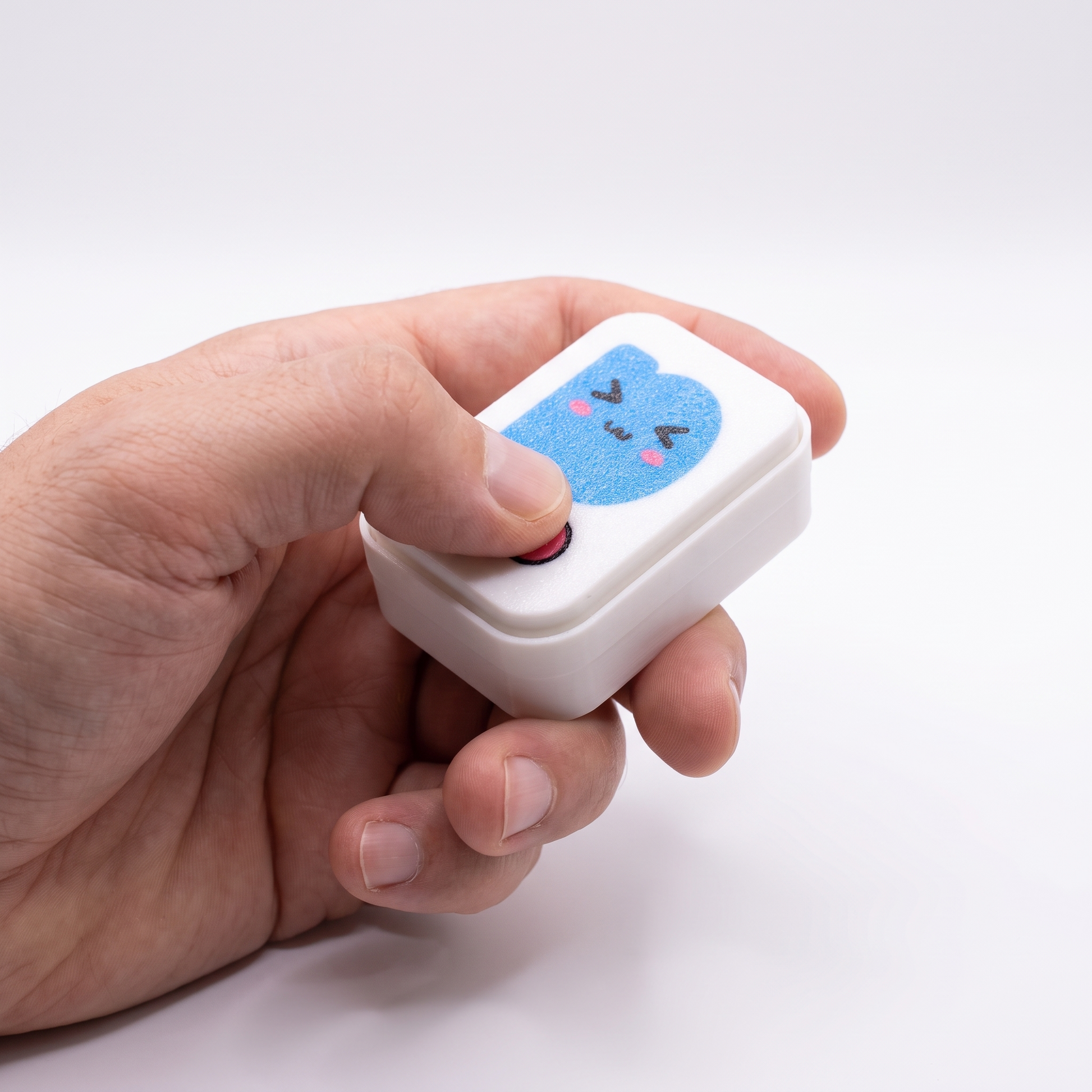 Printies Clicker held in a hand, thumb pressing the pink click button — pocket-sized at 56 × 41 × 24 mm
