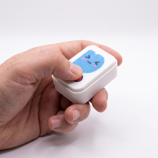 Printies Clicker held in a hand, thumb pressing the pink click button — pocket-sized at 56 × 41 × 24 mm