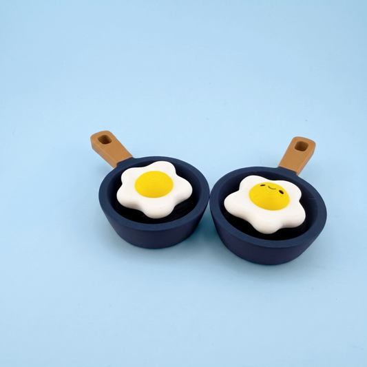 Two 3D printed fried egg fidget clickers in navy frying pans